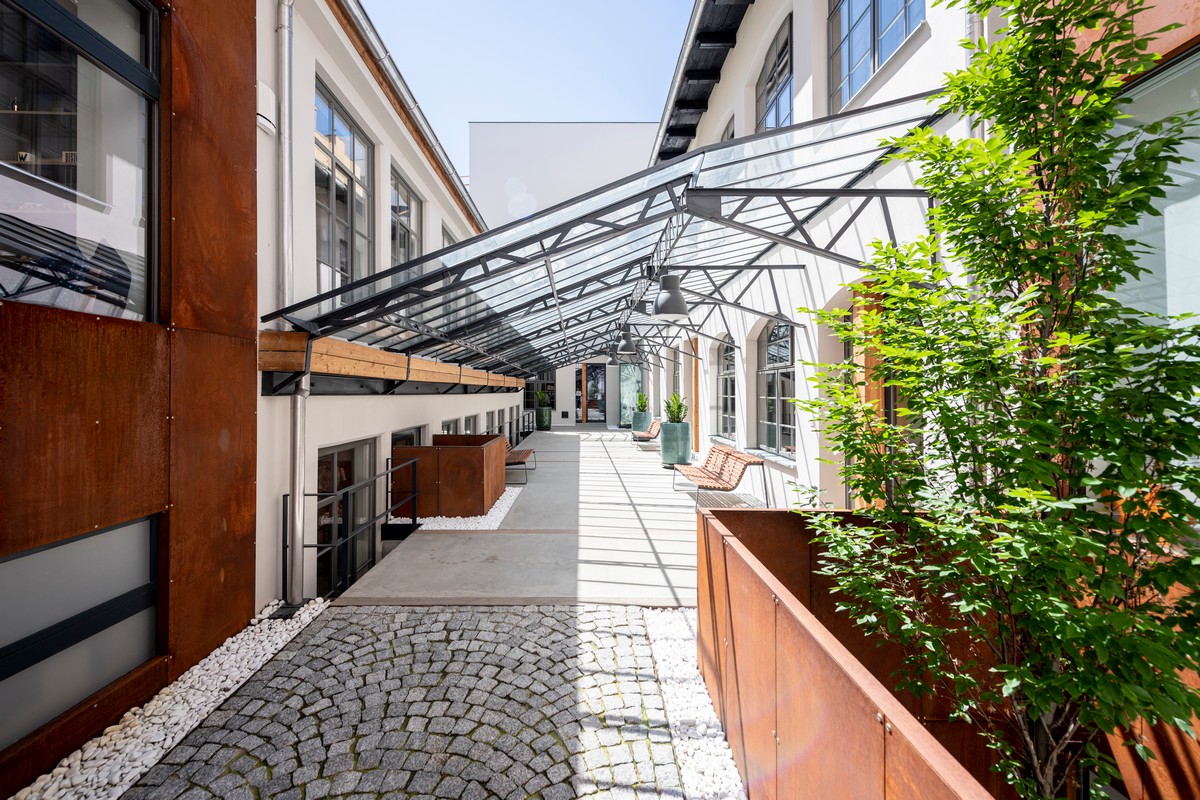 Glass-roofed courtyard walkway between modern white buildings with benches and potted trees.