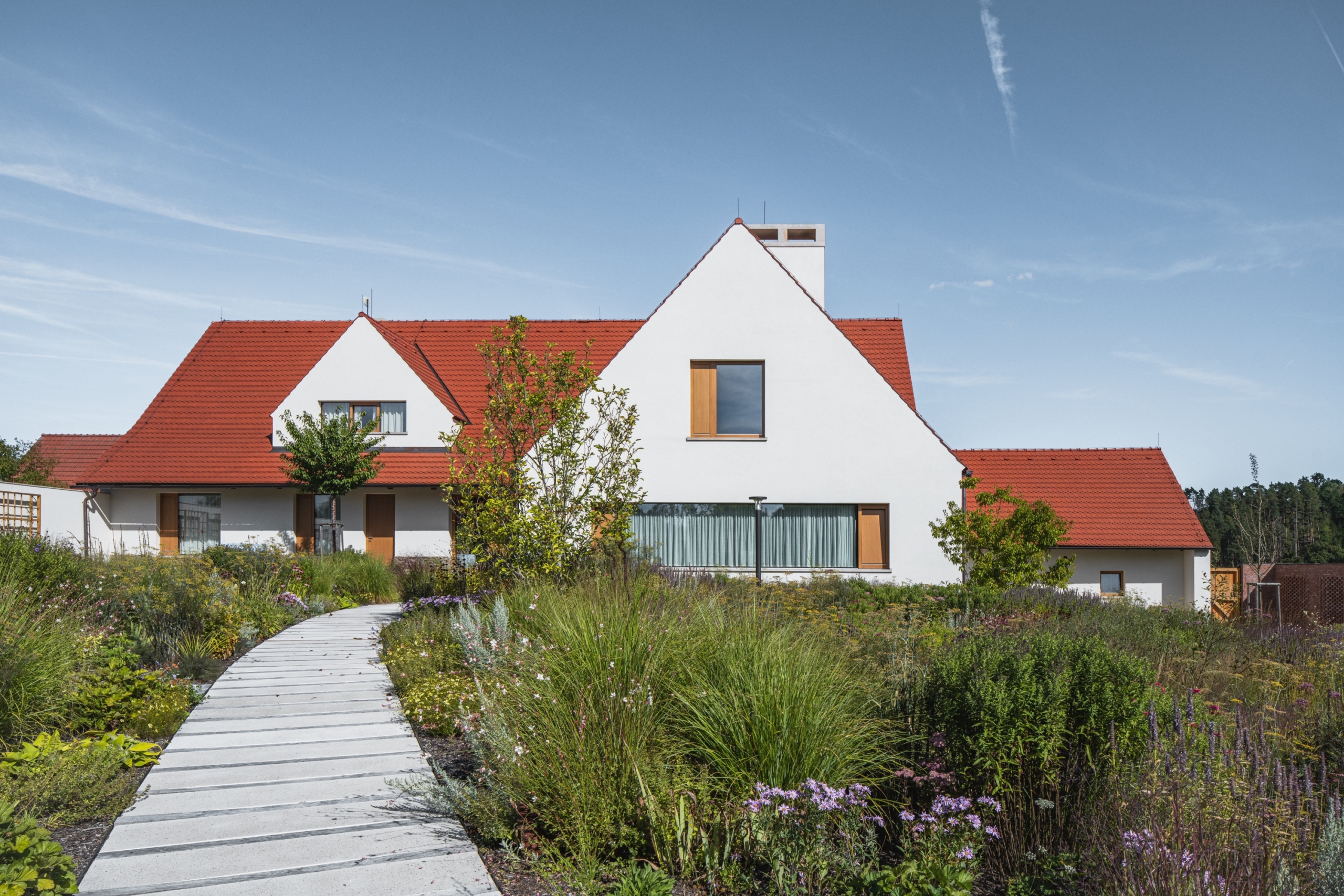 Modern white house with red tiled roof, large windows, and a garden path leading to the entrance under a clear blue sky.